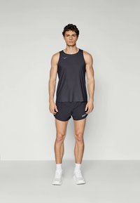 Junger Mann steht, trägt schwarzes ärmelloses Sportshirt und passende Shorts, weiße Socken und Turnschuhe, einfarbiger heller Hintergrund.
