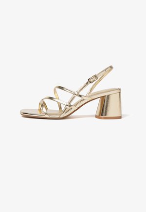 Goldene Riemchensandale mit niedrigem Blockabsatz, verstellbarer Knöchelschnalle und offenem Zehenbereich vor weißem Hintergrund.