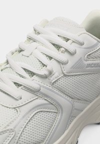 Λευκό αθλητικό παπούτσι με πάνελ από δίχτυ, λευκά κορδόνια και συνθετικές επιστρώσεις, δείχνοντας κοντινή λήψη των λεπτομερειών στο πλάι και στη γλώσσα του παπουτσιού.