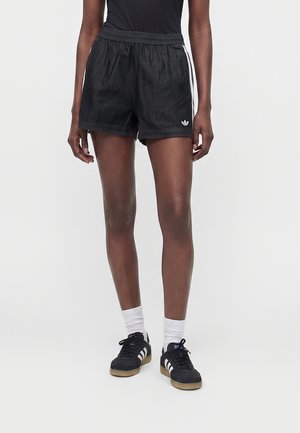 Άτομο που φοράει μαύρα σορτς Adidas με λευκές ρίγες και λογότυπο, μαύρα αθλητικά παπούτσια Adidas με λευκές ρίγες και λευκές κάλτσες, στέκεται σε λευκό φόντο.