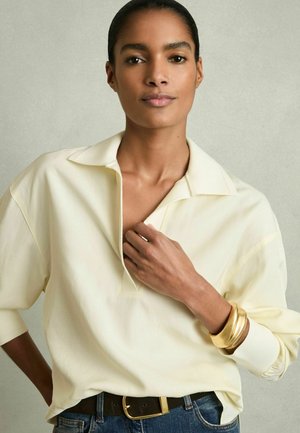 Blusa color crema con colletto e scollatura aperta, maniche lunghe, abbinata a una cinturina scura e bracciali dorati. Tessuto morbido, vestibilità comoda.