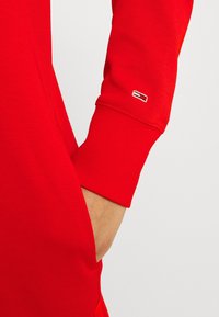 Prenda roja con puños acanalados y un bolsillo lateral, con un pequeño logotipo bordado en la manga. Textura de tejido suave.