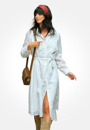 Kobieta w białej sukience z ażurowym wzorem, z przednim rozcięciem, z czerwoną opaską w białe groszki, brązową torebką na ramię i beżowymi kozakami, pozująca na białym tle.