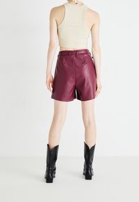 Person som bär en beige ärmlös crop top, höga maroon lädershorts och svarta stövlar som når mitt på vaden, stående mot en vit bakgrund.