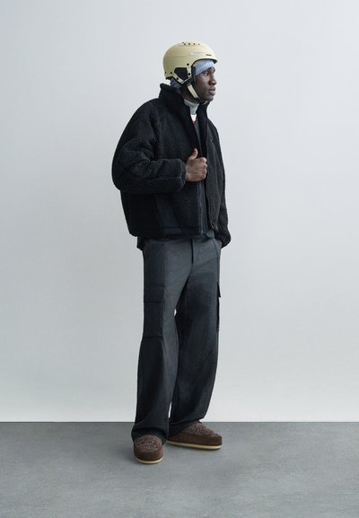 Uomo con giacca in pile nera, pantaloni cargo grigi, pantofole marroni con motivo, casco beige e copricapo blu, in piedi contro un muro semplice.