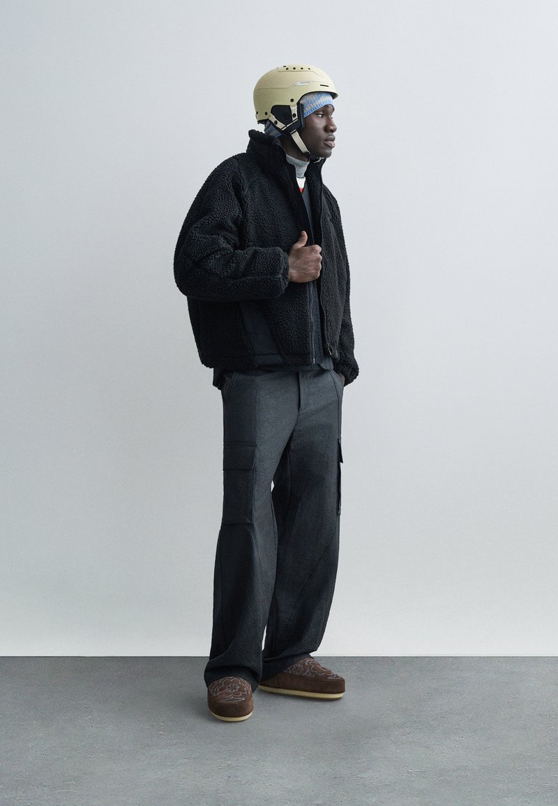 Man wearing black fleece jacket, gray cargo pants, brown patterned slippers, a beige helmet, and a blue head covering, standing against a plain wall.