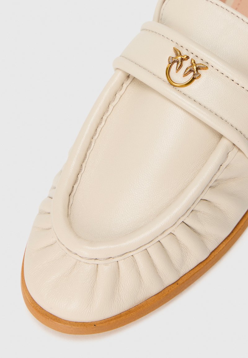 Mocasines de cuero beige con punta redondeada, que presentan detalles fruncidos y un acento dorado de un pájaro en una correa. Suela plana con una textura sutil.