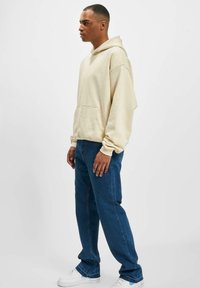 Sudadera crema con bolsillo frontal, corte relajado y capucha ajustable, combinada con jeans azules y zapatillas blancas, presentada de lado.