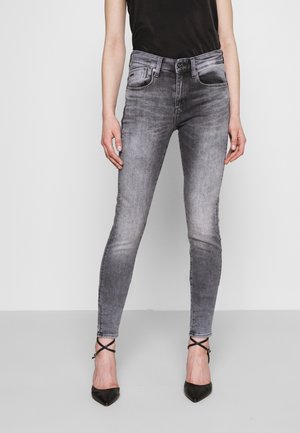 Femme portant un jean skinny gris délavé, des talons noirs à brides et un haut noir sans manches, debout devant un fond clair uni.