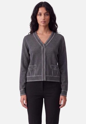 Donna che indossa un cardigan a maniche lunghe grigio scuro con dettagli di cuciture bianche e pantaloni neri, rivolta frontalmente su uno sfondo semplice.