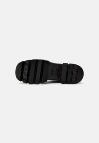 Suela de zapato de goma negra con una superficie texturizada, que presenta surcos y crestas profundas para una mayor tracción. Diseño minimalista con un perfil plano.