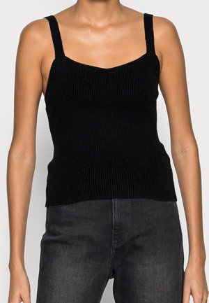 Femme portant un débardeur noir en maille côtelée avec de larges bretelles et un jean gris foncé, montré du cou aux hanches.