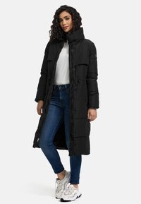 Long black puffer coat with a high collar, front pockets, and a zip closure, worn over a white top and blue jeans, paired with sneakers.