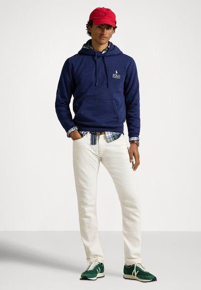 Μπλε σκούρο φούτερ με κουκούλα και μπροστινή τσέπη, φορεμένο πάνω από μπλε καρό πουκάμισο. Λευκό παντελόνι και πράσινα αθλητικά παπούτσια ολοκληρώνουν το σύνολο. Κόκκινο καπέλο για τόνισμα.