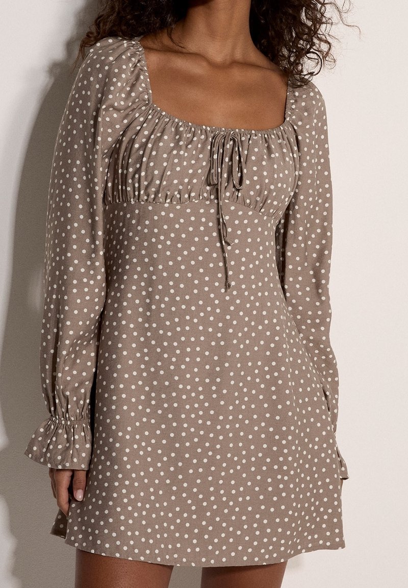Femme portant une robe beige à manches longues avec des pois blancs et un col carré noué au niveau de la poitrine.