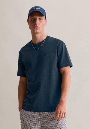 Navy cotton t-shirt with short sleeves, round neckline, and embossed logo detail. Paired with patterned shorts and a cap.