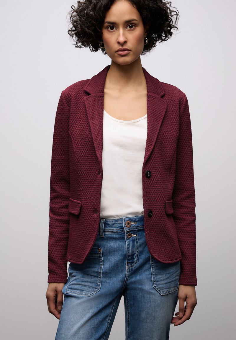 Blazer texturé bordeaux avec deux poches avant et boutons noirs, associé à un haut blanc et un jean en denim bleu avec poches plaquées.