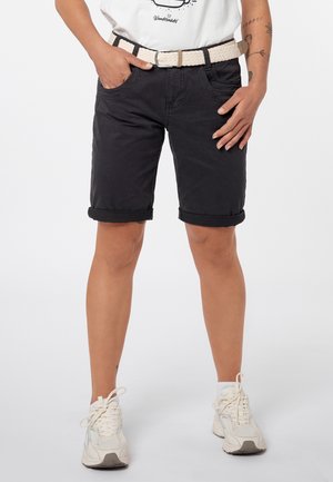 Persoon draagt zwarte opgerolde shorts, witte geweven riem, wit T-shirt met print en witte sneakers, staat met handen bij de zakken.