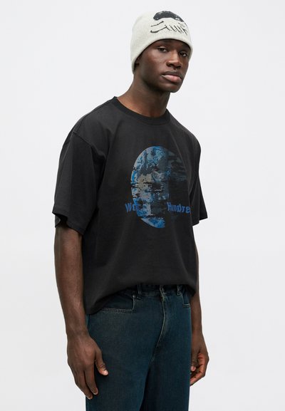 Juodas marškinėliai su mėlyna abstrakčios veido grafika, dėvimi su tamsiai mėlynomis džinsomis. Vyras stovi prieš baltą foną.