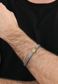Muñeca con pulsera de cadena trenzada de plata que presenta un cierre rectangular de oro, llevada sobre un brazo peludo frente a un fondo de camisa negra.