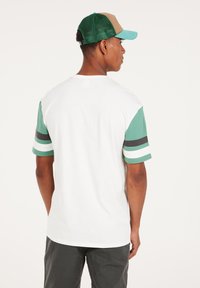 Witte T-shirt met groene en zwarte gestreepte korte mouwen, gecombineerd met een groene mesh pet en een bruine voorkant. Eenvoudig, casual ontwerp.