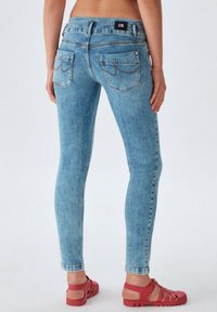 Jean skinny bleu clair avec un lavage moyen, comprenant des poches arrière et une étiquette noire. Porté avec des sandales rouges, montrant une silhouette ajustée.