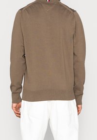 Bruine trui met lange mouwen en geribbelde zoom en manchetten, met een subtiel patroon. Achteraanzicht toont een V-stikdetail nabij de hals.