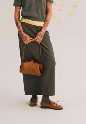 Persona con brazos tatuados vistiendo un top gris y falda, sosteniendo un bolso de cuero marrón, usando mocasines marrones con calcetines oscuros frente a un fondo liso.