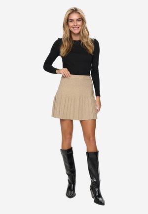 Žena usmievajúca sa, oblečená v čiernom tričku s dlhým rukávom, béžovej plisovanej sukni a čiernych kožených čižmách po kolená, stojaca na bielom pozadí.