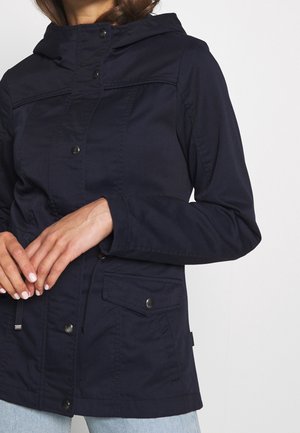 Femme portant une veste à capuche bleu marine avec boutons-pression et poches à rabat, les mains jointes devant, assortie à un jean bleu clair.