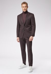 Ciemnobrązowy garnitur o dopasowanym kroju, dopasowany golf i białe sneakersy. Gładka faktura, subtelne wzory na tkaninie oraz zapięcie na dwa guziki.
