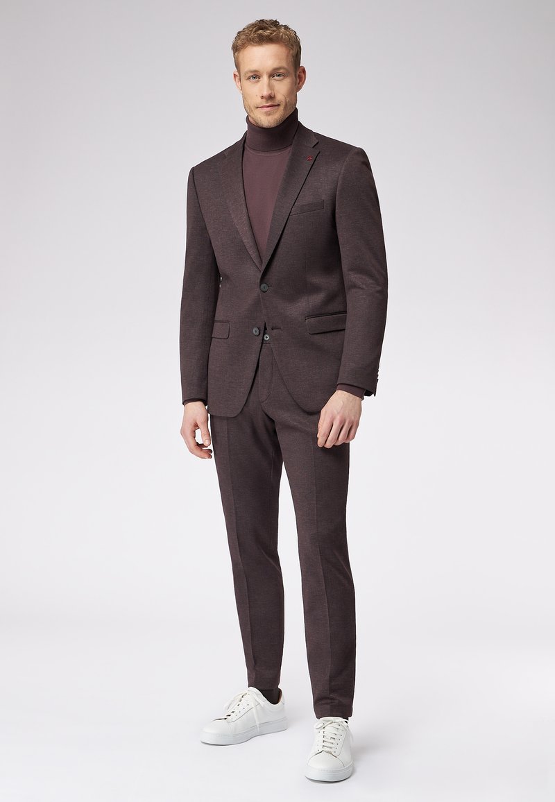 Ciemnobrązowy garnitur o dopasowanym kroju, dopasowany golf i białe sneakersy. Gładka faktura, subtelne wzory na tkaninie oraz zapięcie na dwa guziki.