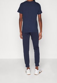 Person steht mit dem Rücken zur Kamera, trägt ein navyblaues Kurzarmhemd, navyblaue Jogginghosen und weiße Sportschuhe vor einem einfarbigen Hintergrund.