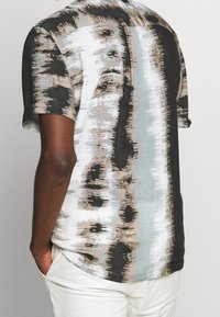 Košile s krátkými rukávy v kombinaci černé, bílé a béžové barvy s abstraktními vzory, s volným střihem a hladkou texturou látky.