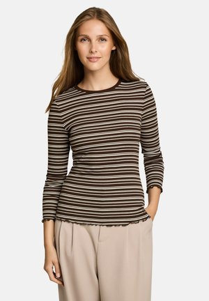 Jeune femme portant une chemise à manches longues rayée marron et blanc et un pantalon beige, une main dans la poche, souriante doucement.