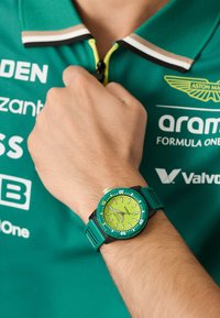 Hodiniek aqua zelená farba s okrúhlym čelom a žltozeleným ciferníkom, ktorý obsahuje biele hodinové značky, čierny rám a textúrovaný remienok.