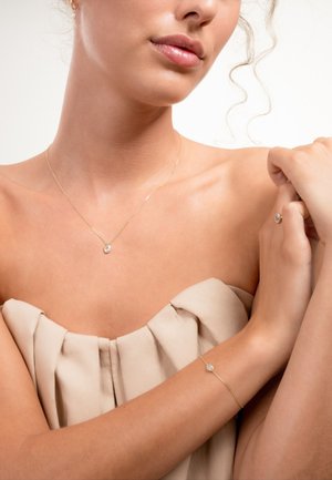 Collier en or avec un pendentif en pierre précieuse claire au centre, associé à un bracelet en or délicat avec une pierre précieuse similaire. Teint de peau lisse, coiffure douce.