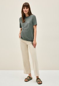 Camiseta de algodón verde oliva con estampado de hojas y diseño gráfico, combinada con pantalones de pierna ancha beige y sandalias verde oscuro.