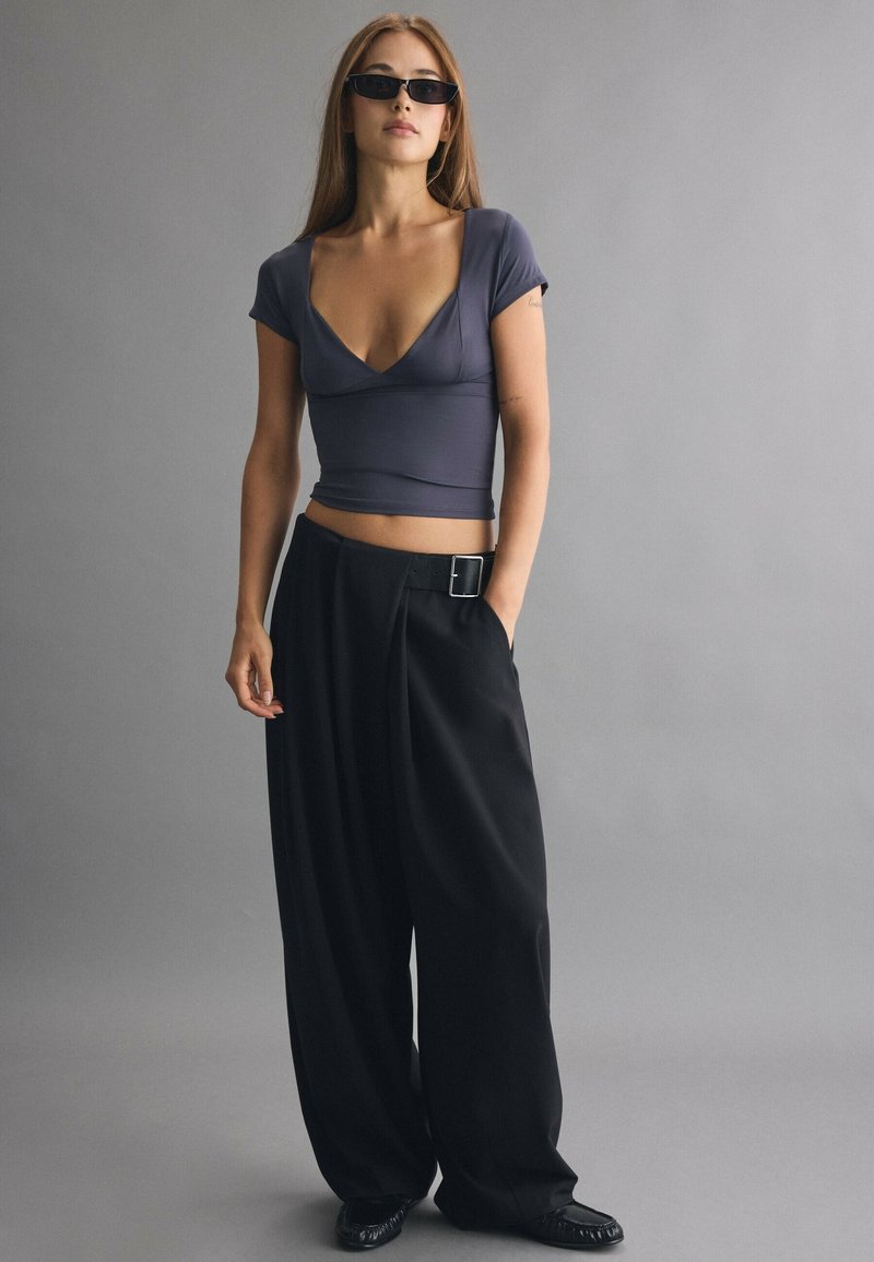 Jeune femme portant des lunettes de soleil foncées, un haut gris cintré à manches courtes, un pantalon large noir avec ceinture et des chaussures noires, debout devant un fond gris uni.