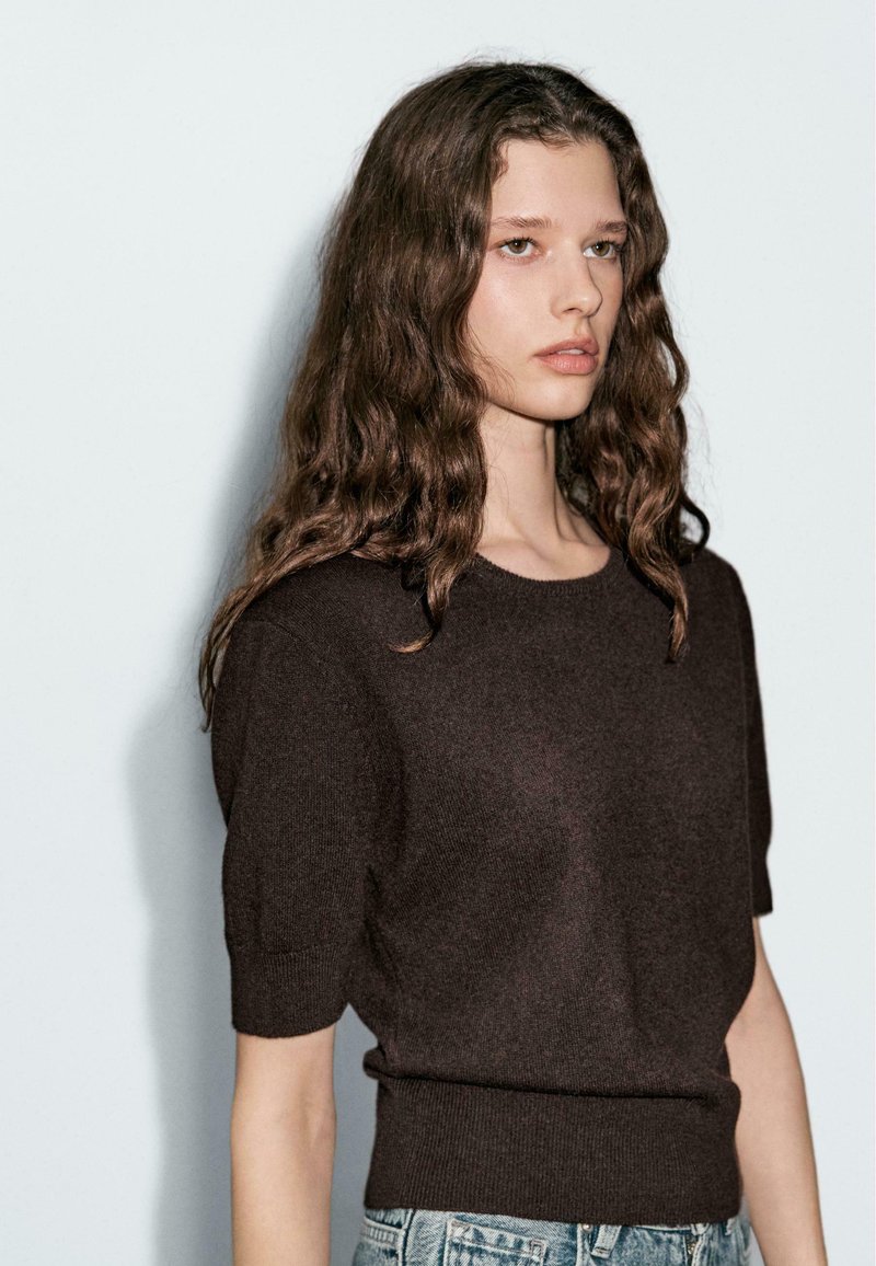 Pull en maille marron à manches courtes avec un col rond, présentant un ourlet côtelé et une texture douce, associé à un jean en denim bleu clair.