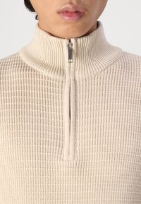 Pull en beige à col montant avec demi-fermeture éclair et motif en maille gaufrée texturée. Doté d'une fermeture éclair en métal et d'un tissu doux.