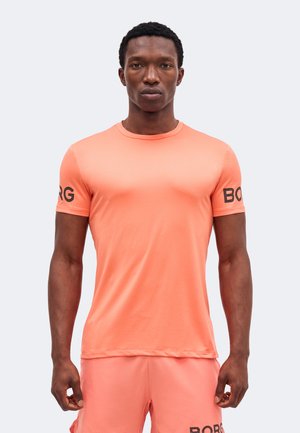 Mężczyzna ubrany w dopasowany koralowy sportowy T-shirt i spodenki z czarnym napisem "BORG" na rękawach i szortach, stojący twarzą do przodu.