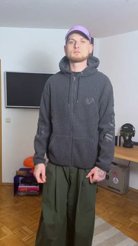 Szara bluza z polaru na suwak z przednimi kieszeniami, zestawiona z luźnymi oliwkowozielonymi spodniami. Całość dopełnia jasnofioletowa czapka.