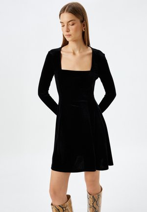 Vestido de terciopelo negro con mangas largas, escote cuadrado y falda acampanada. Llevado con botas marrones de caña alta y con estampado.