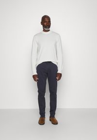 Pull en maille blanche avec un col montant, associé à un pantalon slim bleu marine et des bottines en daim marron. Texture lisse, design simple.