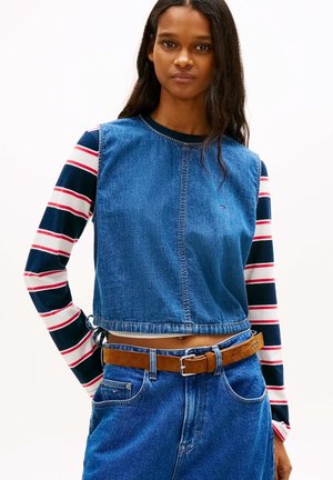 Femme portant un top en jean sans manches par-dessus un t-shirt à manches longues rayé bleu marine, blanc et rouge, avec un jean bleu et une ceinture marron, debout devant un fond blanc.