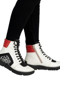 Białe botki do kostki z czarnymi podeszwami, czerwonymi wyściełanymi kołnierzami, czarnymi sznurowadłami, bocznymi zamkami błyskawicznymi oraz czarno-białymi panelami w wzory, noszone z czarnymi spodniami.