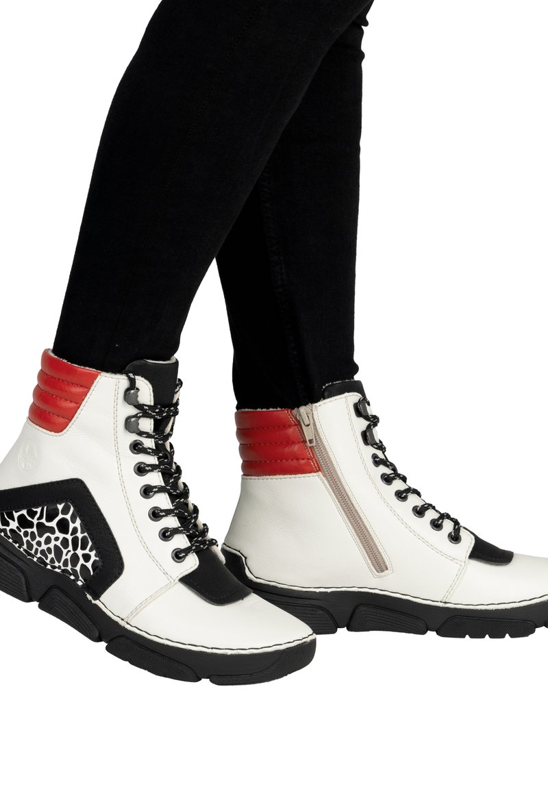 Białe botki do kostki z czarnymi podeszwami, czerwonymi wyściełanymi kołnierzami, czarnymi sznurowadłami, bocznymi zamkami błyskawicznymi oraz czarno-białymi panelami w wzory, noszone z czarnymi spodniami.