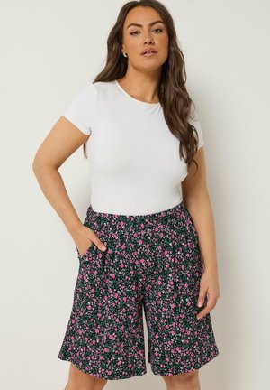 Kobieta o długich brązowych włosach, ubrana w biały krótki top z krótkim rękawem i czarne spodenki z drobnym różowym kwiatowym wzorem, ręce w kieszeniach.