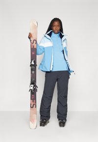 Ski jakke i lyseblå med hvide striber, parret med mørke skibukser. Holder en snowboard med bindinger, iført sorte skistøvler.
